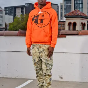 Man in an orange hoodie and camouflage pants standing outdoors.