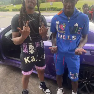Two young men posing in front of a purple car, wearing casual streetwear.
