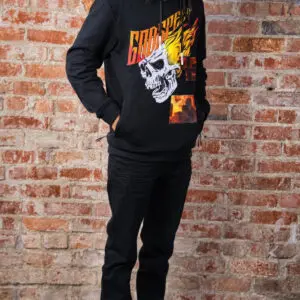 Person wearing a black hoodie with a flaming skull graphic and black pants against a brick wall.