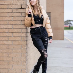 Woman in a tan jacket and ripped black jeans leaning against a brick wall.